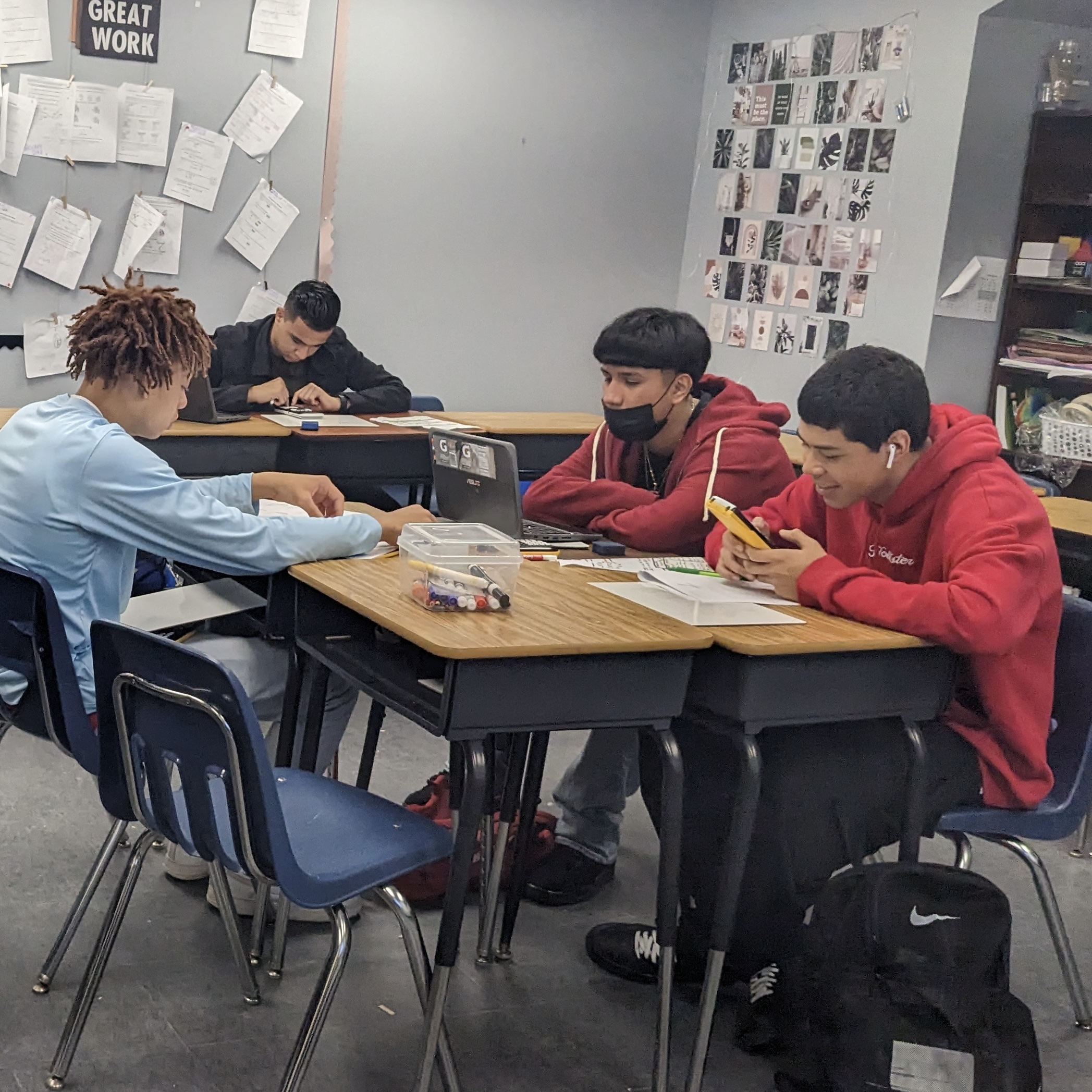 Four students sitting at desks using Chromebooks, graphing calculators, and whiteboards in a classroom setting.
