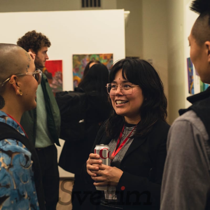 Lidia laughing mid-conversation at the Our Light Unbound opening night, with exhibition artwork on the gallery walls behind her.