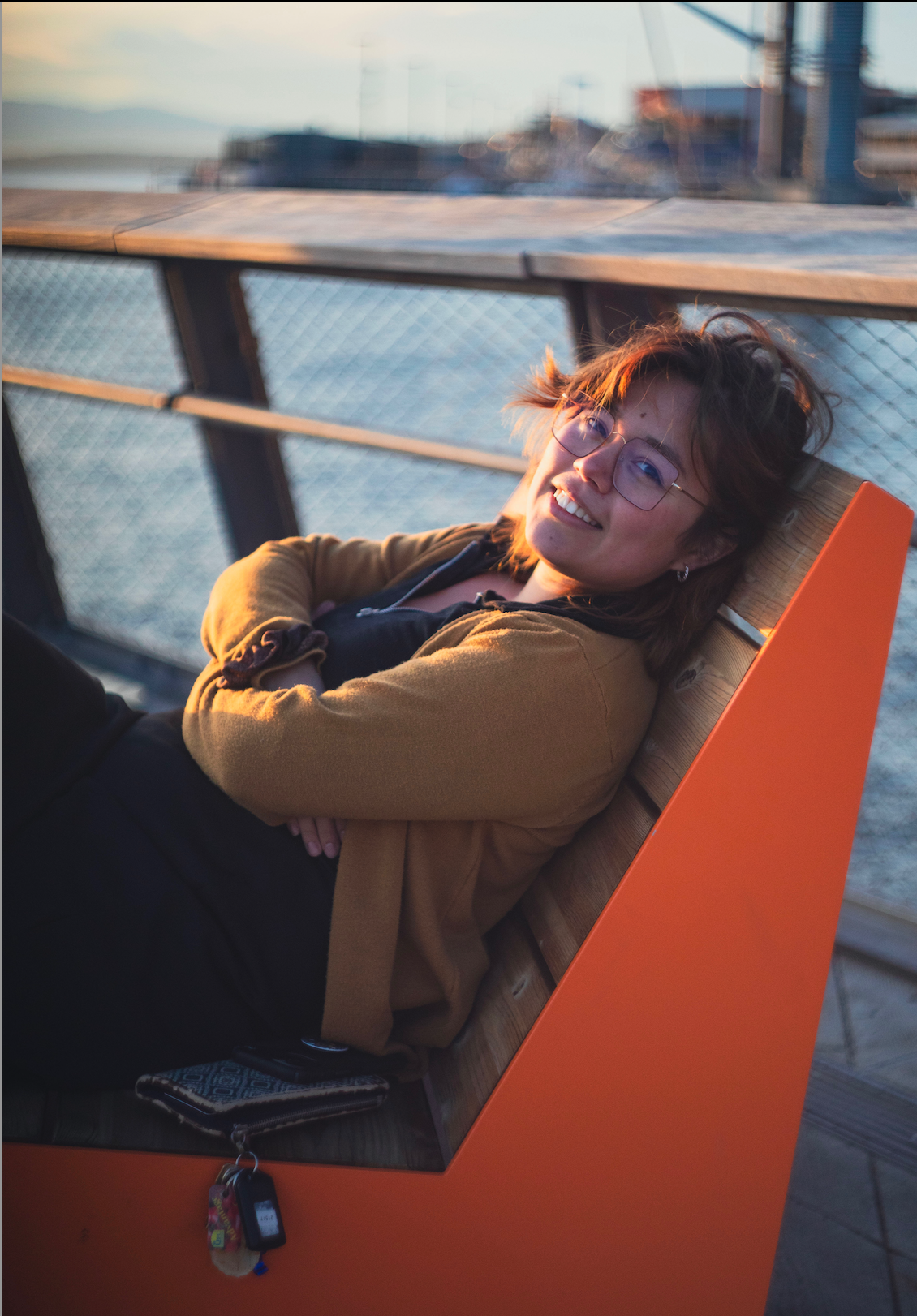 Lidia smiles, wearing a mustard yellow cardigan and reclining on a bright orange park bench.
