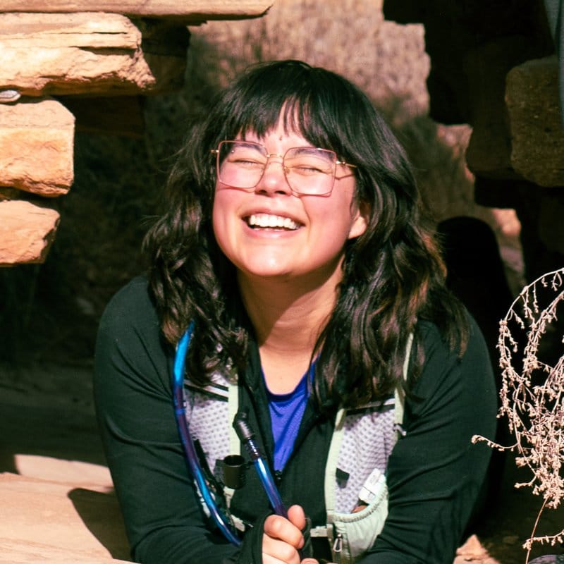 Lidia grins while peeking through a stone opening at a desert ruins site.