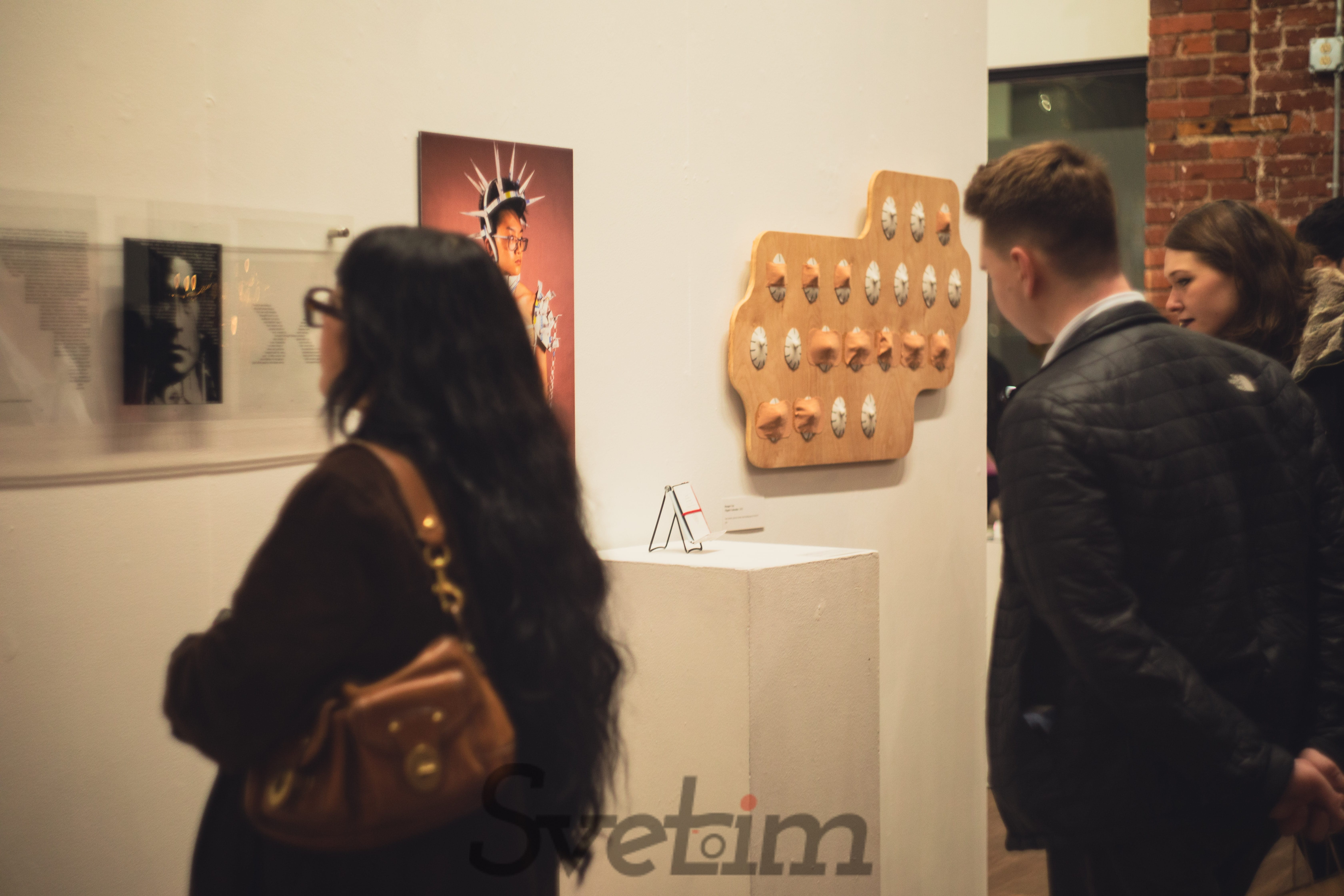 Gallery wall showing a mix of photography, sculpture, and mixed media art — small printed art labels visible beside each piece