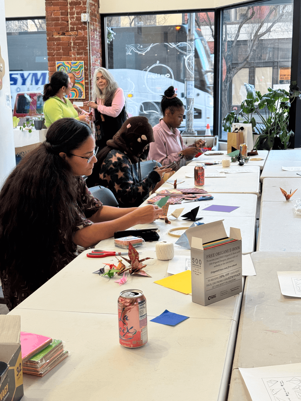 Participants folding origami cranes at a long table during the origami mobile workshop, with colorful paper and completed cranes visible