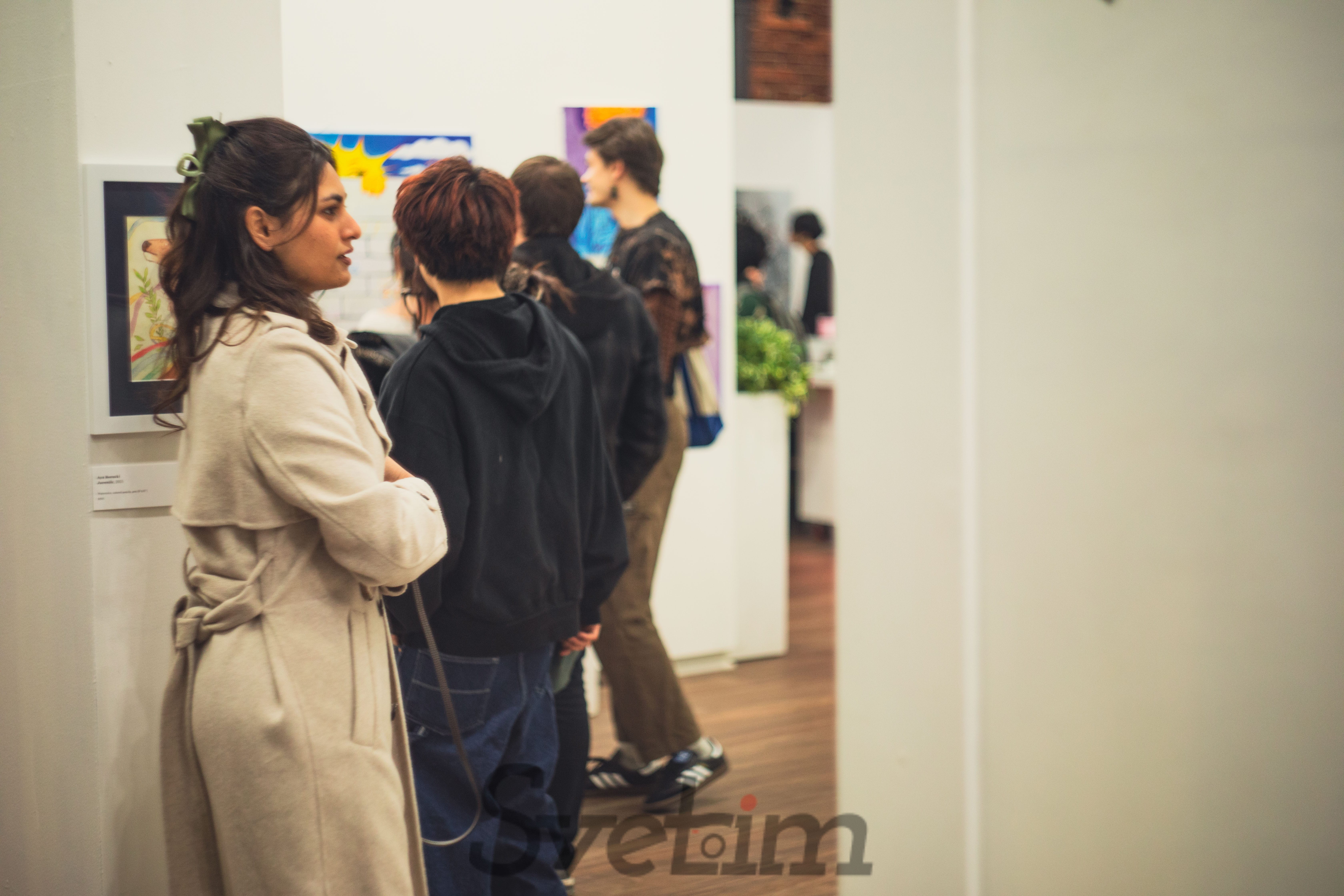 A visitor pausing to examine a framed artwork on the gallery wall, with other visitors browsing deeper in the space