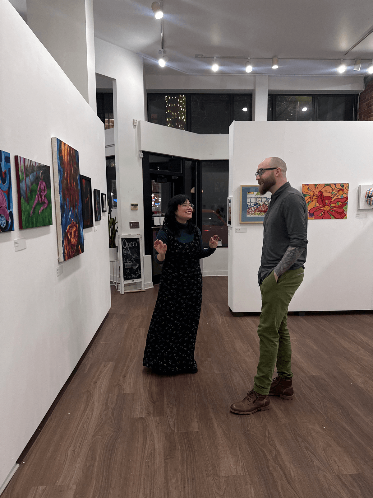 A curator talking with a visitor about the artwork on display, standing in the gallery surrounded by colorful paintings
